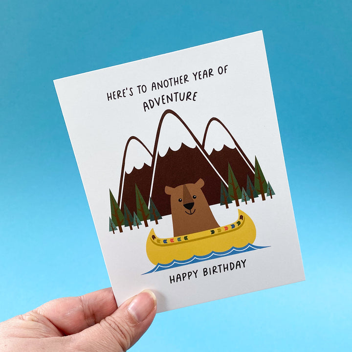 Birthday card with bear in a kayak and mountain illustration, held against a blue background.