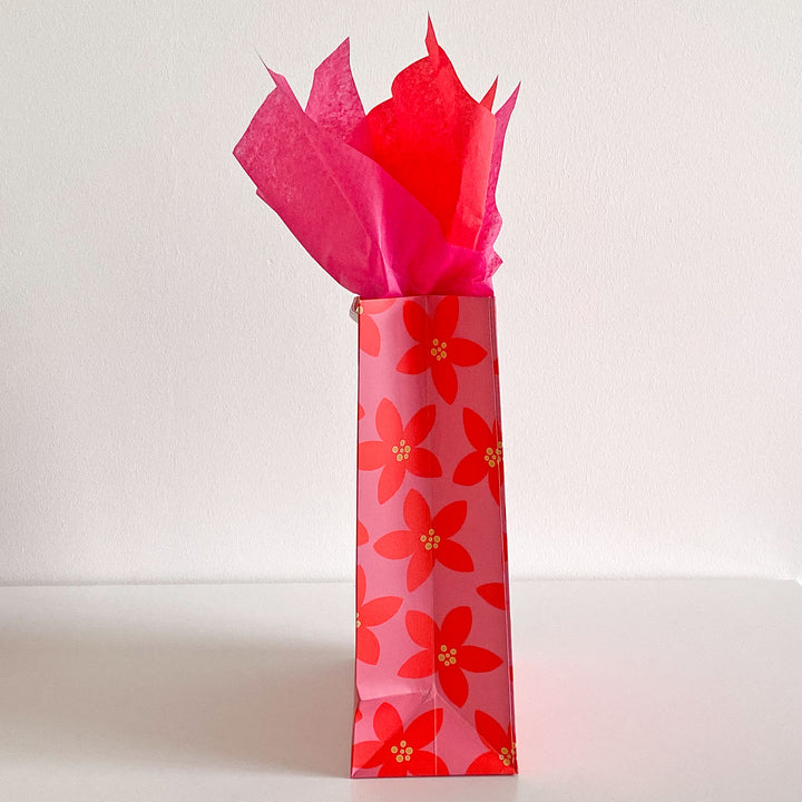 Side view of a red floral-patterned gift bag with pink tissue paper on a white background
