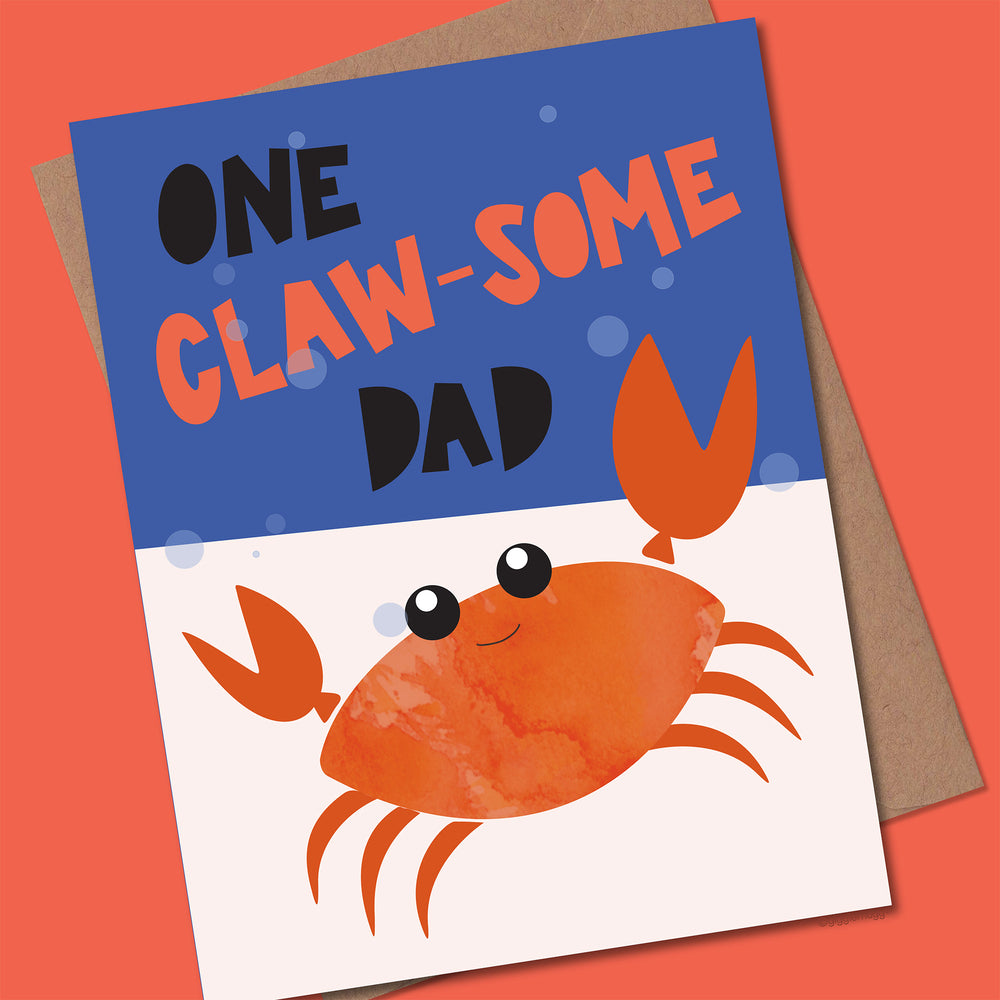 Greeting card with a crab illustration and text 'One Claw-some Dad' with a Kraft envelope on a red background.