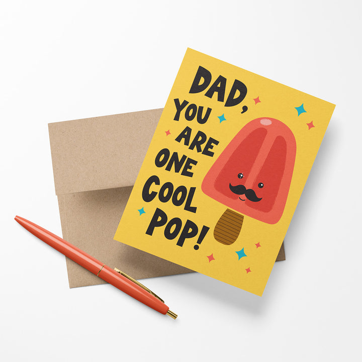 Illustrated greeting card for Dad with a red popsicle character and text that says, "Dad, you are one cool pop!" shown with a Kraft envelope and pen.