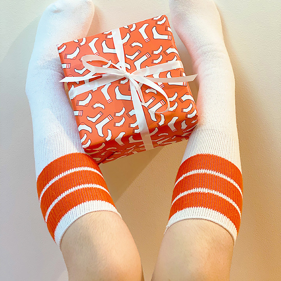 Two feet in tube socks holding a gift wrapped in fun socks wrapping paper tied with white ribbon by ©Gigglemugg