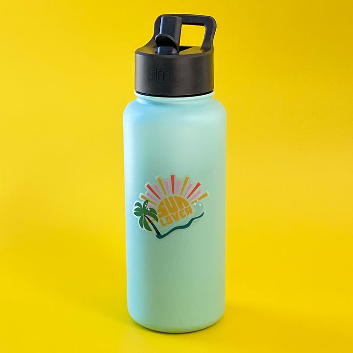 Light blue water bottle with black lid on a yellow background. Water bottle features a die-cut vinyl "Sun Lover" sticker.