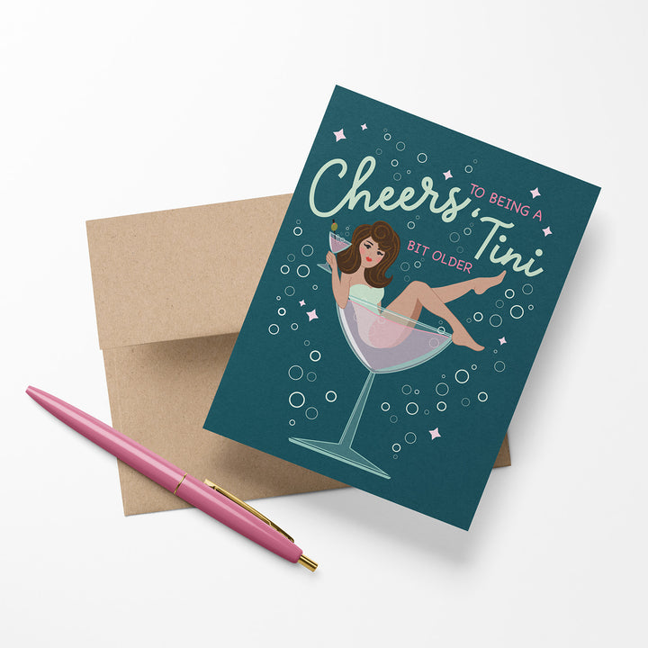 Greeting card with illustration of a woman in a martini glass and humorous text, "Cheers to being a tini bit older." Shown with envelope and pen.