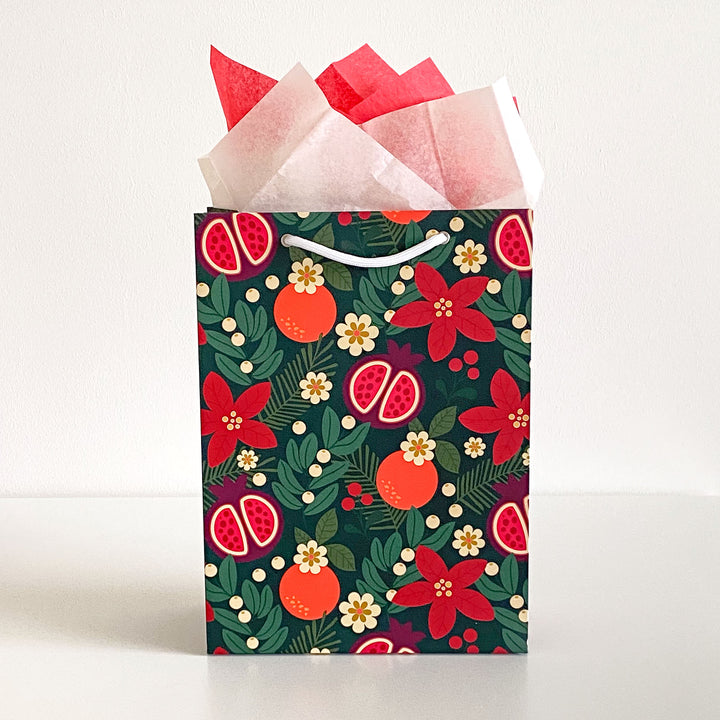 A gift bag featuring a holiday fruit and floral pattern with red and cream colored tissue paper