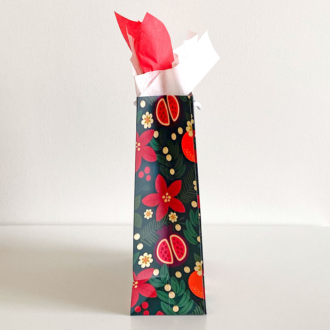 A gift bag featuring a holiday fruit and floral pattern with red and cream colored tissue paper. Shown in side view.