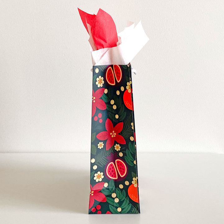 A gift bag featuring a holiday fruit and floral pattern with red and cream colored tissue paper. Shown in side view.
