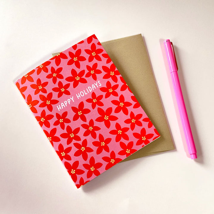 A holiday card with a floral pattern sitting on a kraft envelope, shown next to a pink pen.